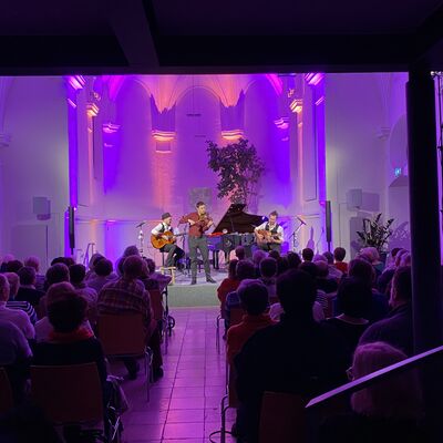 Blick auf die lila und blau ausgeleuchtete Bühne der Spitalkirche. Auf der Bühne spielt das Trio Cobario auf zwei Gitarren und einer Geige.