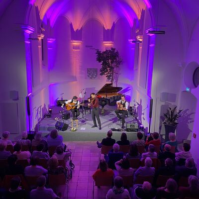 Blick von der Empore auf die lila und blau ausgeleuchtete Bühne der Spitalkirche. Auf der Bühne spielt das Trio Cobario auf zwei Gitarren und einer Geige.