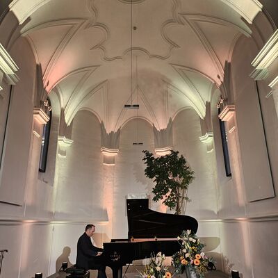 Der Pianist Christian Seibert sitzt an an dem geöffneten Flügel auf der Bühne der Schwandorfer Spitalkirche. Vor dem Flügel sind zwei Vasen mit Blumenarrangements.