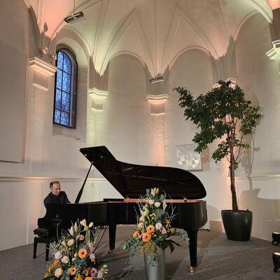Der Pianist Christian Seibert sitzt an an dem geöffneten Flügel auf der Bühne der Schwandorfer Spitalkirche. Vor dem Flügel sind zwei Vasen mit Blumenarrangements.