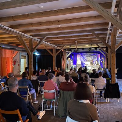 Foto von einem Konzert im Sperlstadel. Es ist ein alter Stall mit Holzdecke und Holzstützen. Menschen sitzen mit Abstand vor der Bühne und hören der Musikgruppe The Jules Band zu.
