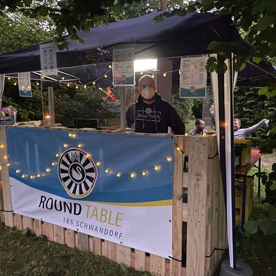 Foto von einer Bar. Überdacht mit einem Pavillon. Hinter der Bar steht ein Mann mit Maske hinter einer Plexiglasschutzscheibe. An der Bar hängt ein Werbebanner auf dem Round Table Schwandorf steht.
