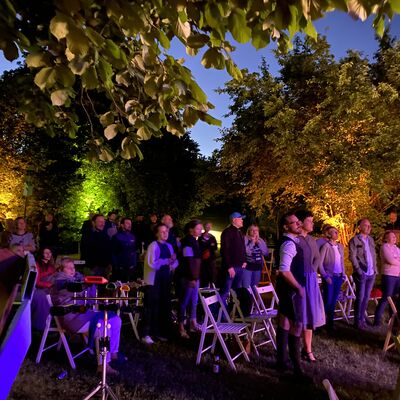 Foto von Menschen, die vor der Open-Air Bühne am Abend im Park des Oberpfälzer Künstlerhauses stehen. Sie sind in lila Licht angeleuchtet, man sieht viele Bäume.
