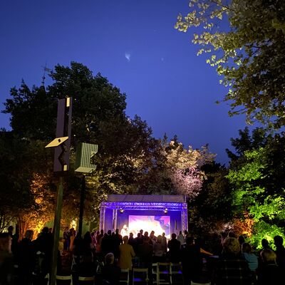 Foto vom Park des Oberpfälzer Künstlerhauses mit Open-Air Bühne. Es dämmert, die Bäume sind beleuchtet. Menschen stehen vor einer Open-Air Bühne und hören der Musikgruppe Loisach Marci zu.
