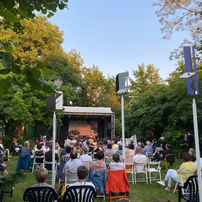 Foto mit sommerlicher Atmosphäre. Menschen sitzen auf Stühlen im Skultpurenpark des Oberpfälzer Künstlerhauses und schauen auf eine Open-Air Bühne. Auf der Bühne spielen die Singer-Songwriter Ami Warning & Matthew Austin.