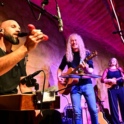 Drei Musiker spielen umgeben von roter Beleuchtung auf der Bühne. Ein Mann im linken Bild singt und spielt ein Perkussionsinstrument. Rechts daneben spielt ein weiterer Mann die Gitarre. Im Hintergrund spielt eine Frau Ukulele.