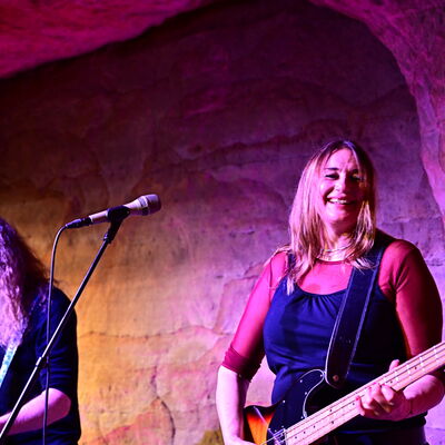 Ein Mann spielt die Gitarre und neben ihm spielt eine Frau lächelnd den Bass. Die Personen sind vom warmen Licht auf der Bühne umgeben.