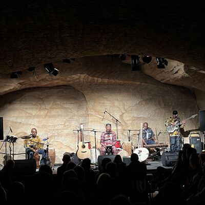 Vier Personen sind auf der Bühne und davon spielen jeweils zwei akustische Instrumente. Die anderen zwei spielen Gitarre und Keyboard. Vereinzelte Personen aus dem Publikum sind zu sehen.