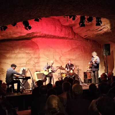 Vier Musiker sind auf der Bühne. Drei Männer sitzend an einem Keyboard, mit einer Gitarre und an einem Schlagzeug. Die vierte Person spielt im Stehen die Gitarre.