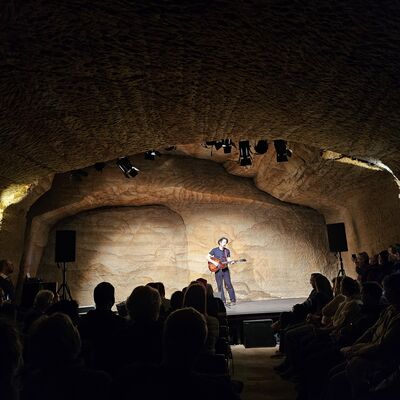 Ein Musiker spielt auf der Bühne Gitarre und singt vor dem sitzenden Publikum.