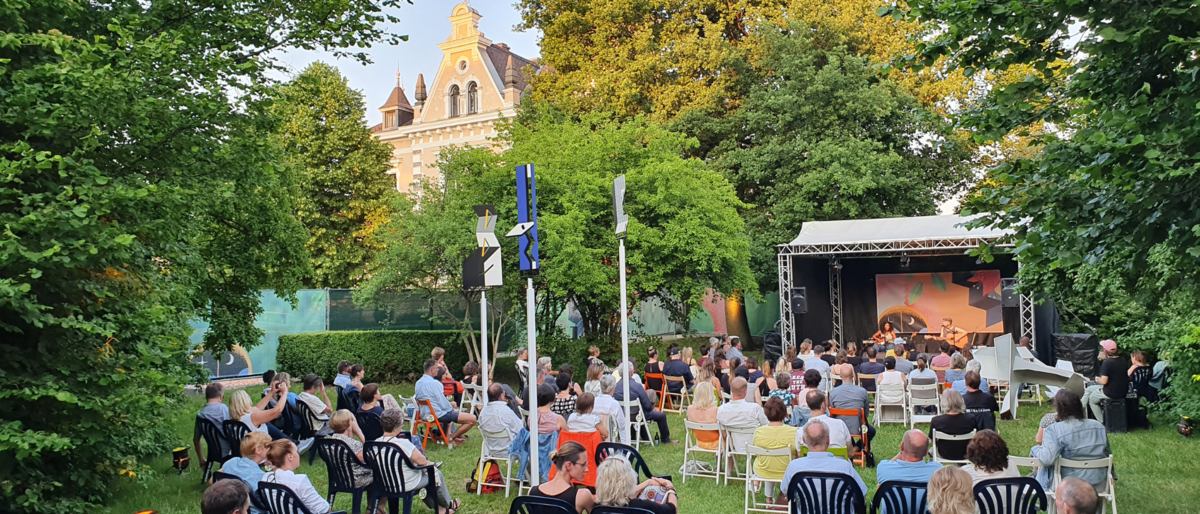 Foto von Menschen, die auf Sthlen im Park vor einer Open-Air Bhne sitzen. Zwischen den Sthlen ist Abstand. Im Hintergrund ist das Oberpflzer Knstlerhaus. Frhsommerliches Ambiente.