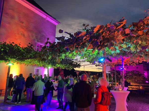 Menschen sind auf dem bunt erleuchteten gläsernen Tanzboden in der Kirchengasse versammelt. Auch die Platanen um den Tanzboden und die Hausfassade sind bunt beleuchtet.
