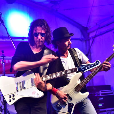 Bild vergrößern: Foto zweier Gitarristen, die auf einer Bühne performen. Die Hintergrundbeleuchtung ist blau.