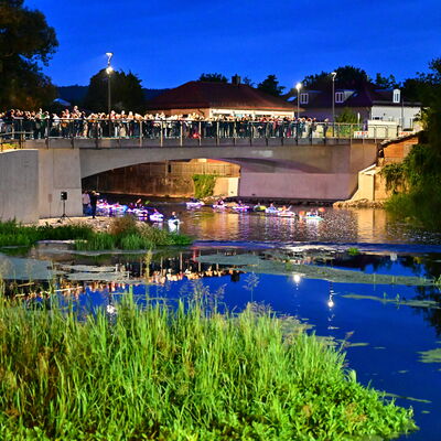 Bild vergrößern: Der Kanuclub Schwandorf paddelt mit bunt beleuchteten Kanus auf der nächtlichen Naab. Viele Menschen schauen von der Brücke aus zu.
