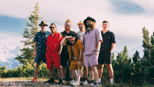 Bild vergr&ouml;&szlig;ern: Die sieben Mitglieder der Band Loamsiada. Hinter ihnen sind B&auml;ume und ein Bergpanorama zu sehen.