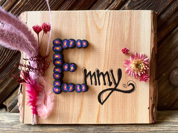 Bild vergr&ouml;&szlig;ern: Auf eine Holzscheibe ist k&uuml;nstlerisch der Name "Emmy" geschrieben. Daneben sind rosa Bl&uuml;ten geklebt.