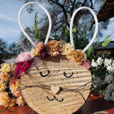 Auf eine Holzscheibe ist ein Hasengesicht gemalt. Er hat Ohren aus wei&szlig;en Pfeiffenputzern und einen Blumenkranz