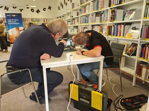Bild vergr&ouml;&szlig;ern: zwei M&auml;nner sitzen an einem Tisch und untersuchen einen elektronischen Gegenstand.