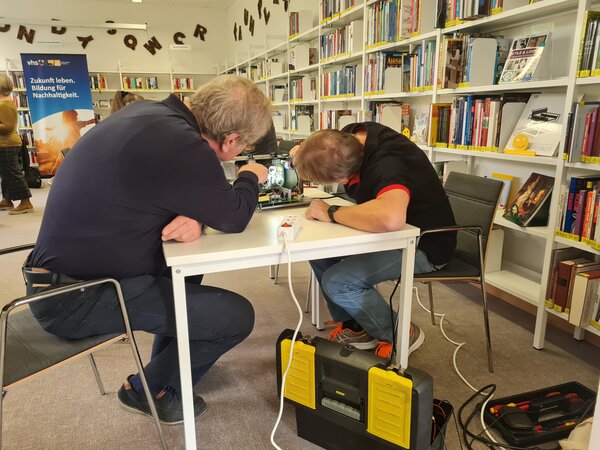 Bild vergr&ouml;&szlig;ern: zwei M&auml;nner sitzen an einem Tisch und untersuchen einen elektronischen Gegenstand.