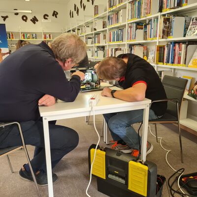 zwei M&auml;nner sitzen an einem Tisch und untersuchen einen elektronischen Gegenstand.