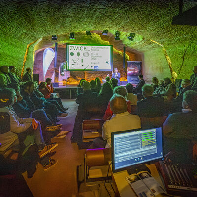Das Publikum sitzt im gr&uuml;nbeleuchteten Felsenkeller und schaut auf eine Leinwand.