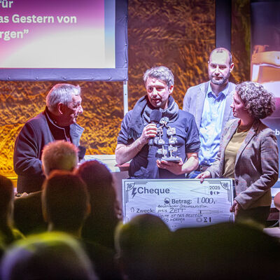 Im Vordergrund des Bildes befinden sich drei M&auml;nner und eine Frau. Der Mann, der in der Mitte steht, spricht in ein Mikrophon und das Publikum h&ouml;rt ihm zu. Es ist der Regisseur des Films "Heute ist das gestern von morgen". Dies findet im bunt beleuchteten Felsenkeller statt. Er bekommt einen Scheck f&uuml;r den Bayerischen Dokumentarfilmpreis ZETT &uuml;berreicht.