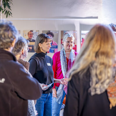 Im Zentrum des Bildes steht eine Frau, die etwas erz&auml;hlt und die Leute, die um sie herumstehen, h&ouml;ren ihr aufmerksam zu.