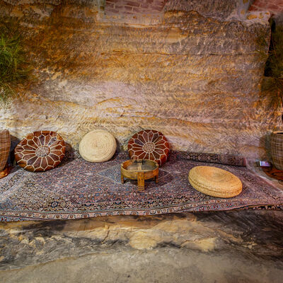 Im Zentrum des Bildes, liegt ein dunkelfarbiger Teppich auf den Boden des Felsenkellers. Auf dem Teppich liegen zwei braunfarbige Lederkissen und zwei hellbraune Strohkissen. Neben dem Teppich, auf der linken Seite, steht eine Pflanze und eine Strohlampe.