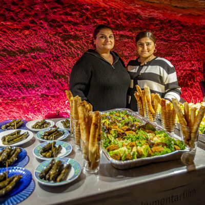 Im rot beleuchteten Felsenkeller, ist ein Buffet aufgebaut und dahinter stehen zwei Frauen, die das Essen verkaufen.