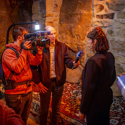 Im Zentrum des Bildes befinden sich zwei M&auml;nner und eine Frau. Der eine Mann hat ein Mikrophon in der Hand und interviewt damit die Frau, w&auml;hrenddessen der andere Mann das Gespr&auml;ch mit einer Filmkamera aufnimmt..