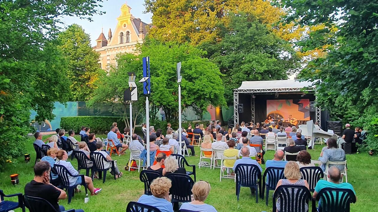 Bild vergrößern: Foto der Bühne der Come-Together Konzertreihe im Park bei der Kebbelvilla. Die Open-Air-Bühne wird von Bäumen umrahmt. Im Vordergrund sitzen Zuschauer:innen.