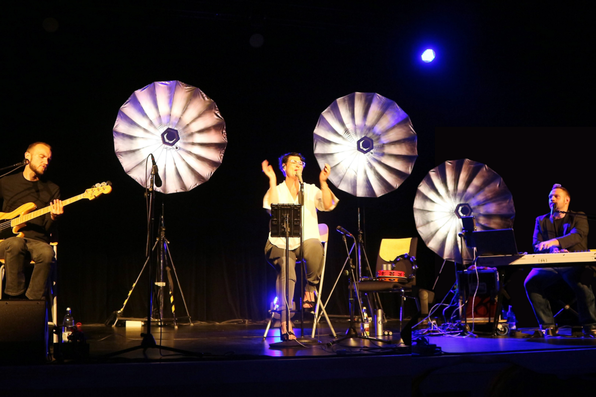 Foto der Band Soulfood unplugged, die auf einer dunklen Bühne performt. Im Hintergrund sind helle Schirmlampen zu sehen.