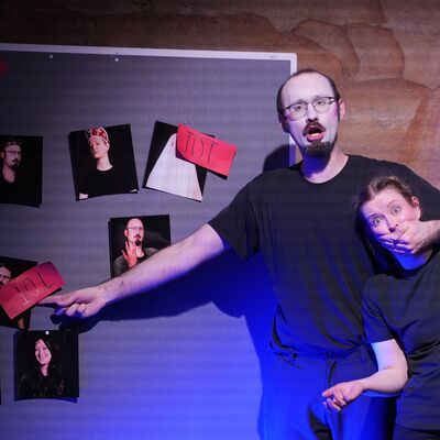 Ein Schauspieler mit wei&szlig; geschminktem Gesicht deutet auf eine Pinnwand, auf die mehrere Fotos gepinnt sind. Mit der anderen Hand verdeckt er einer Schauspielerin mit fragender Pose den Mund.