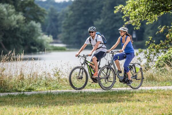 Bild vergrößern: Foto von zwei Fahrradfahrenden, die auf einem Weg vor einem Wasserkörper fahren. Die Sonne scheint.
