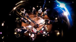Die Musiker:innen des Anarchist Brass Collective stehen im Kreis und spielen ihre Instrumente. Das Bild ist aus der Vogelperspektive mit einer Fish-Eye-Linse aufgenommen. 