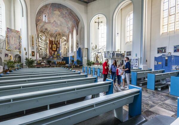 Eine Familie mit einem Fhrer bei der Kirchenfhrung in der Kreuzbergkirche. Die Bnke in der Kirche sind hellblau und im Hintergrund sind die groen Fenster und der prchtige Altar zu sehen.
