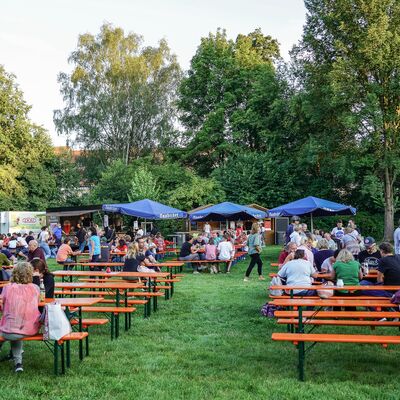 Bild vergrößern: Foto von Bierbänken und Tischen sowie Sonnenschirmen im Stadtpark.