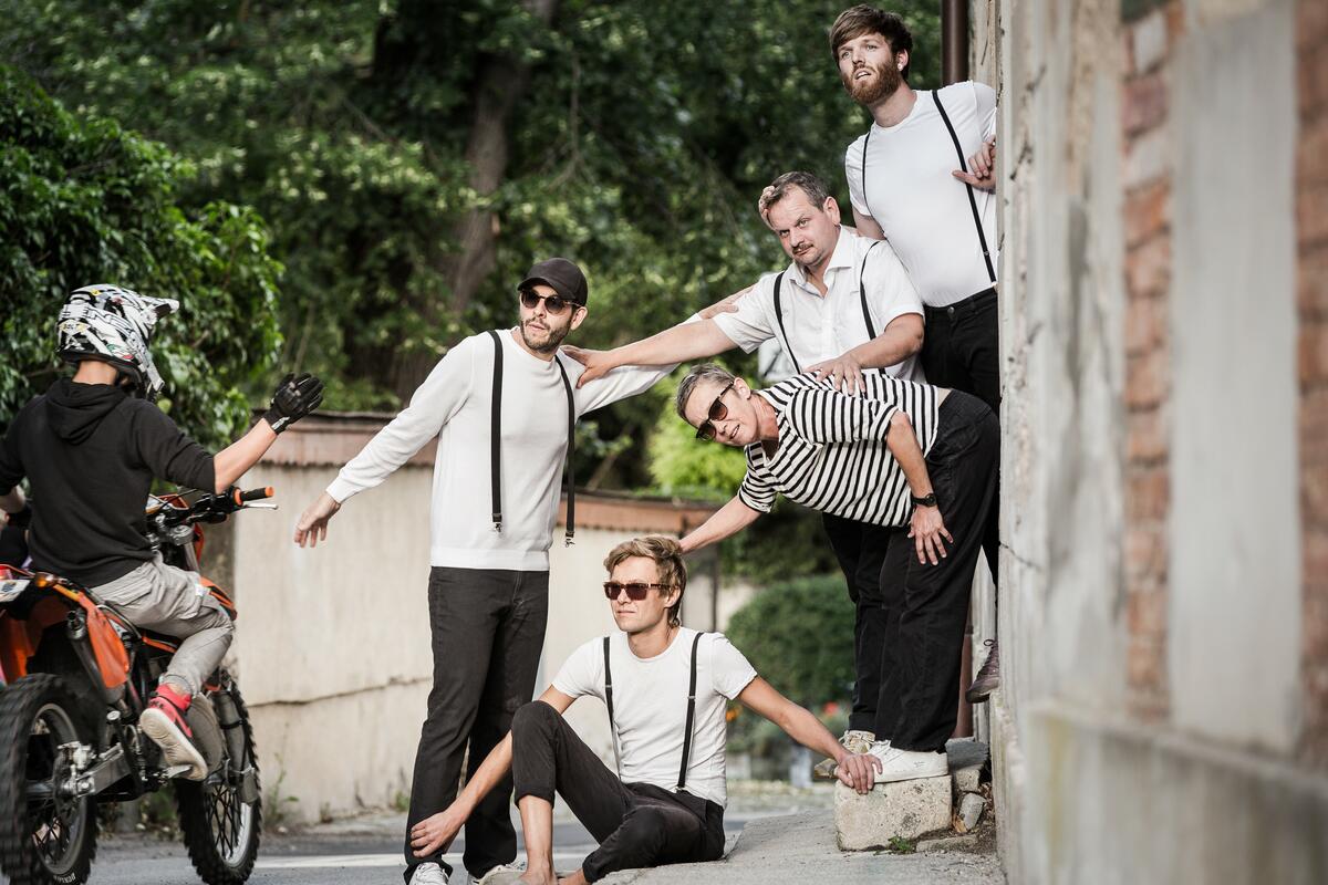 Foto der Gruppe 5/8erl in Ehr'n. Die fünf Bandmitglieder posieren in einem Hauseingang und schauen auf die Straße. Sie tragen schwarz-weiße Kleidung und teilweise Hosenträger und Sonnenbrillen.