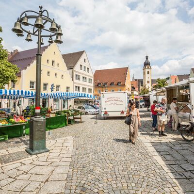 Schwandorfer Bauernmarkt an einem sonnigen Tag auf dem Marktplatz. Es sind Personen, die einkaufen und im Hintergrund die Jakobskirche zu sehen.
