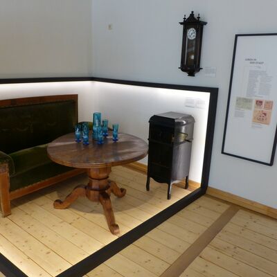 Bild vergrößern: Foto eines Ausstellungsraumes, in dem ein historisches Wohnzimmer dargestellt wird: Ein Sofa aus Samt, ein Säulentisch, ein Grammophon, eine Wanduhr und ein blauer Glaskrug mit sechs Gläsern.