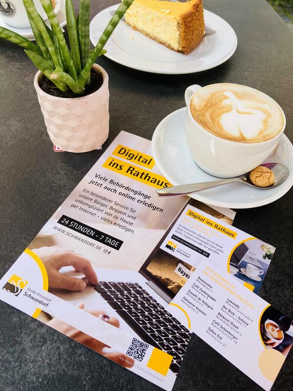 Bild vergrößern: Ein Blumentopf, eine Tasse Cappuccino, ein Stück Kuchen und ein Plakat.