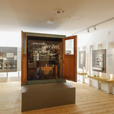 Bild vergrößern: Foto der  mechanischen Turmuhr aus der Kirche St. Georg in Klardorf. Sie steht in einer Vitrine mit weißem Schriftzug: Gebraucht der Zeit, sie geht so schnell von hinnen. Johann Wolfgang von Goethe (1749-1832)